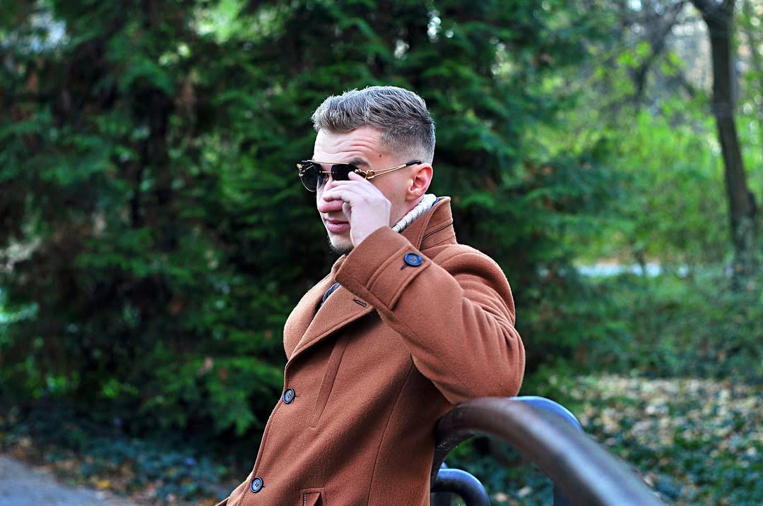 Jakub Kasperek w parku, brązowy płaszcz, okulary przeciwsłoneczne